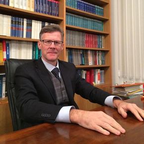 Uomo in abito seduto a una scrivania di legno in un ufficio, con scaffali pieni di libri alle sue spalle