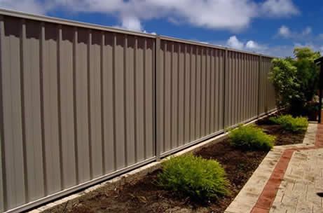 A tall, light-colored metal privacy fence runs along a garden bed with small green shrubs and a stone-paved walkway.
