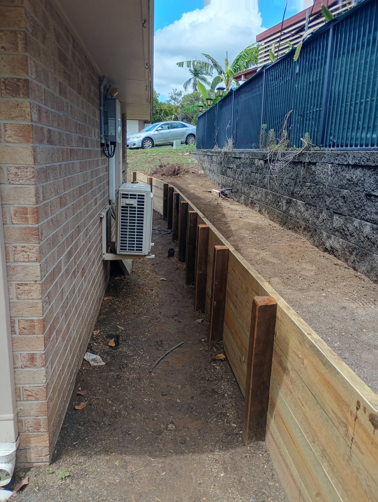 A narrow dirt path between a brick house with an air conditioning unit and a new timber sleeper retaining wall.