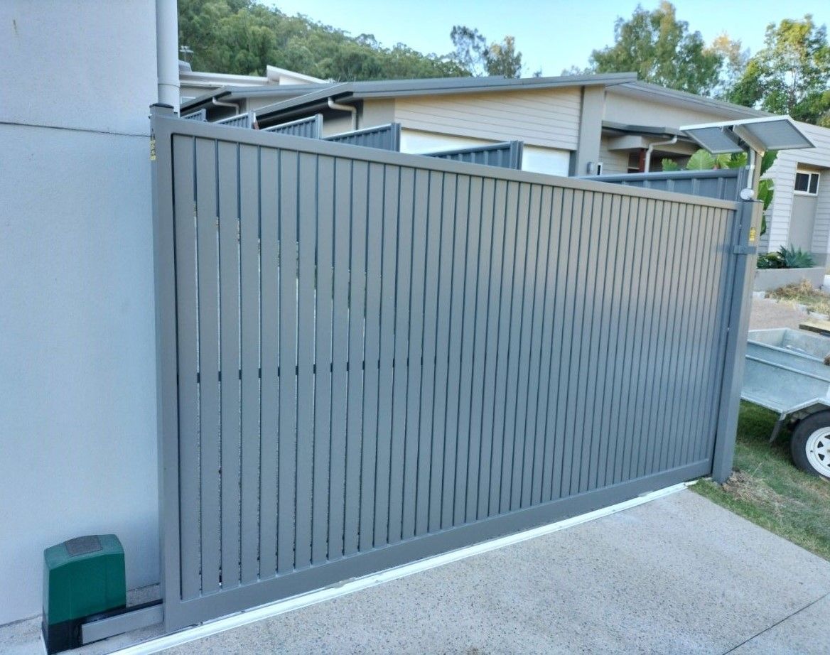A gray, vertical-slat sliding driveway gate with an automatic motor at its base, installed on a residential concrete path.