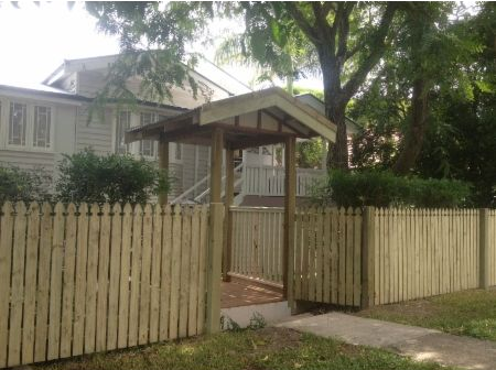 A house with light gray horizontal siding, partially obscured by a wooden picket fence and a covered gateway in the yard.
