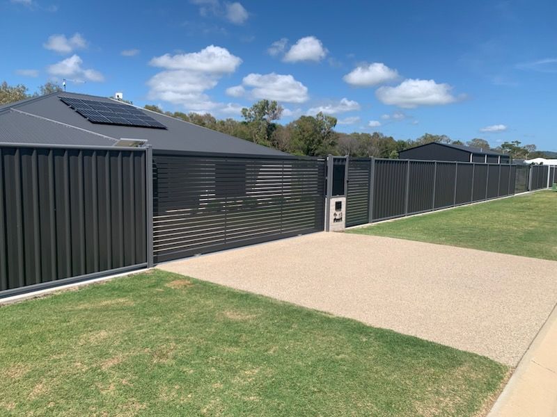 A modern house featuring a dark metal privacy fence and sliding gate next to a concrete driveway and green lawn.