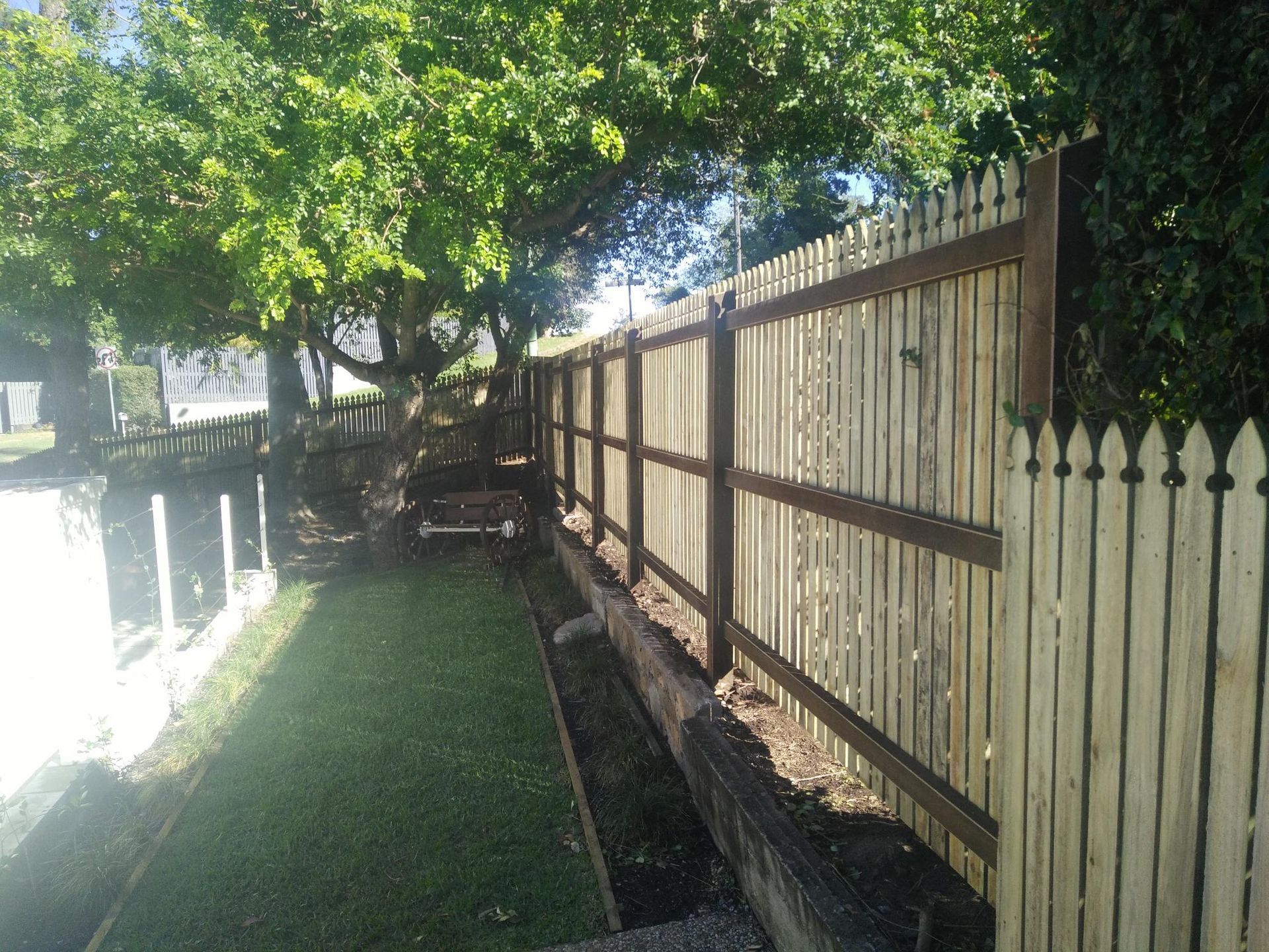 A wooden picket fence borders a strip of grass and a low garden bed, with trees casting shadows over the area.
