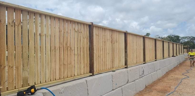 A wooden fence built atop a concrete block retaining wall outdoors under a cloudy sky.