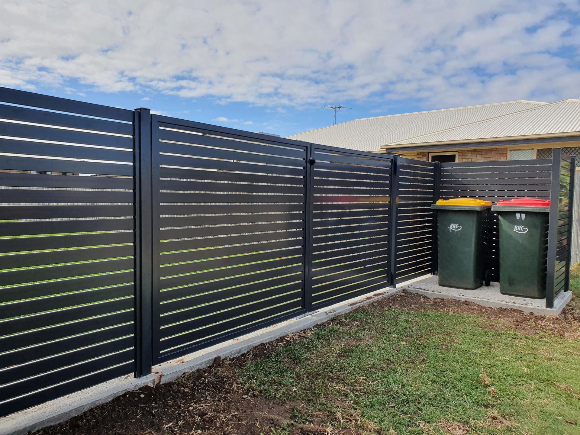 A modern black horizontal slat metal fence borders a backyard with two wheelie bins stored in a corner.