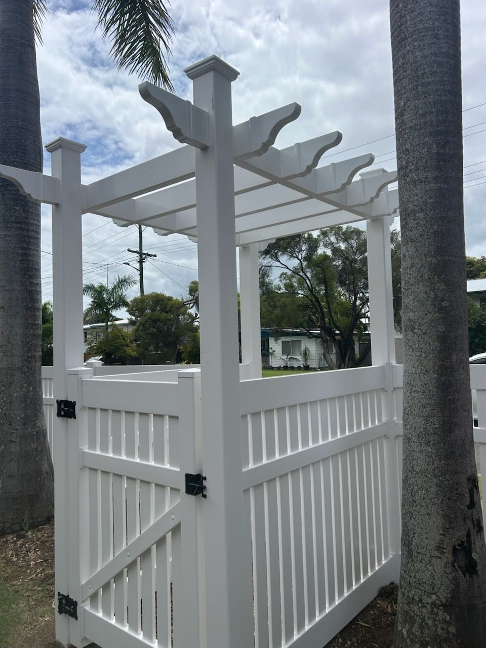 White wooden gate and pergola structure. Trees frame the view, sky background.