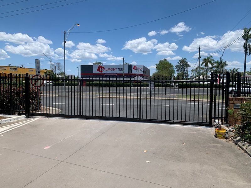 A black metal sliding gate blocks a driveway, with a paved road and a large warehouse building in the background.