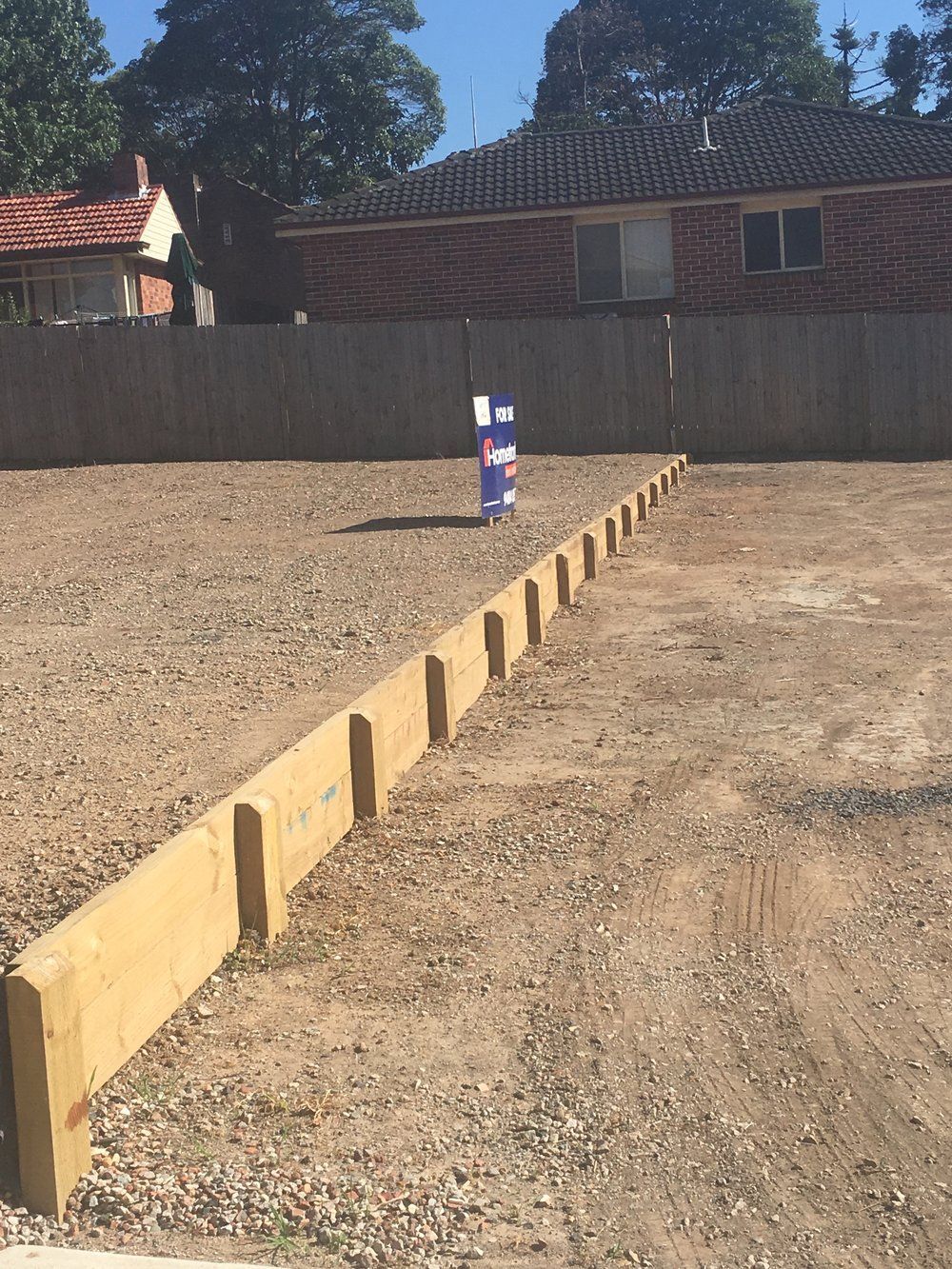 A wooden retaining wall separates two sections of a cleared, gravel-filled residential lot under a clear blue sky.