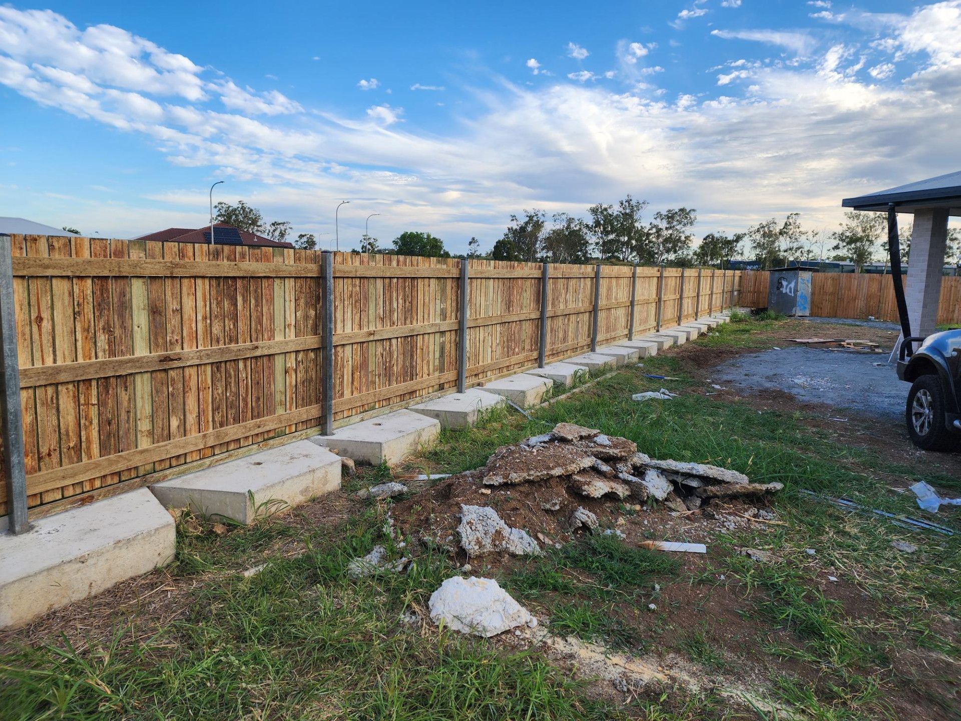 A wooden fence with metal posts, supported by a row of concrete blocks along the base, set in a grassy yard under a blue sky.