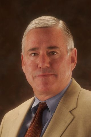 Man in light tan suit, blue shirt, and patterned tie, smiling slightly.