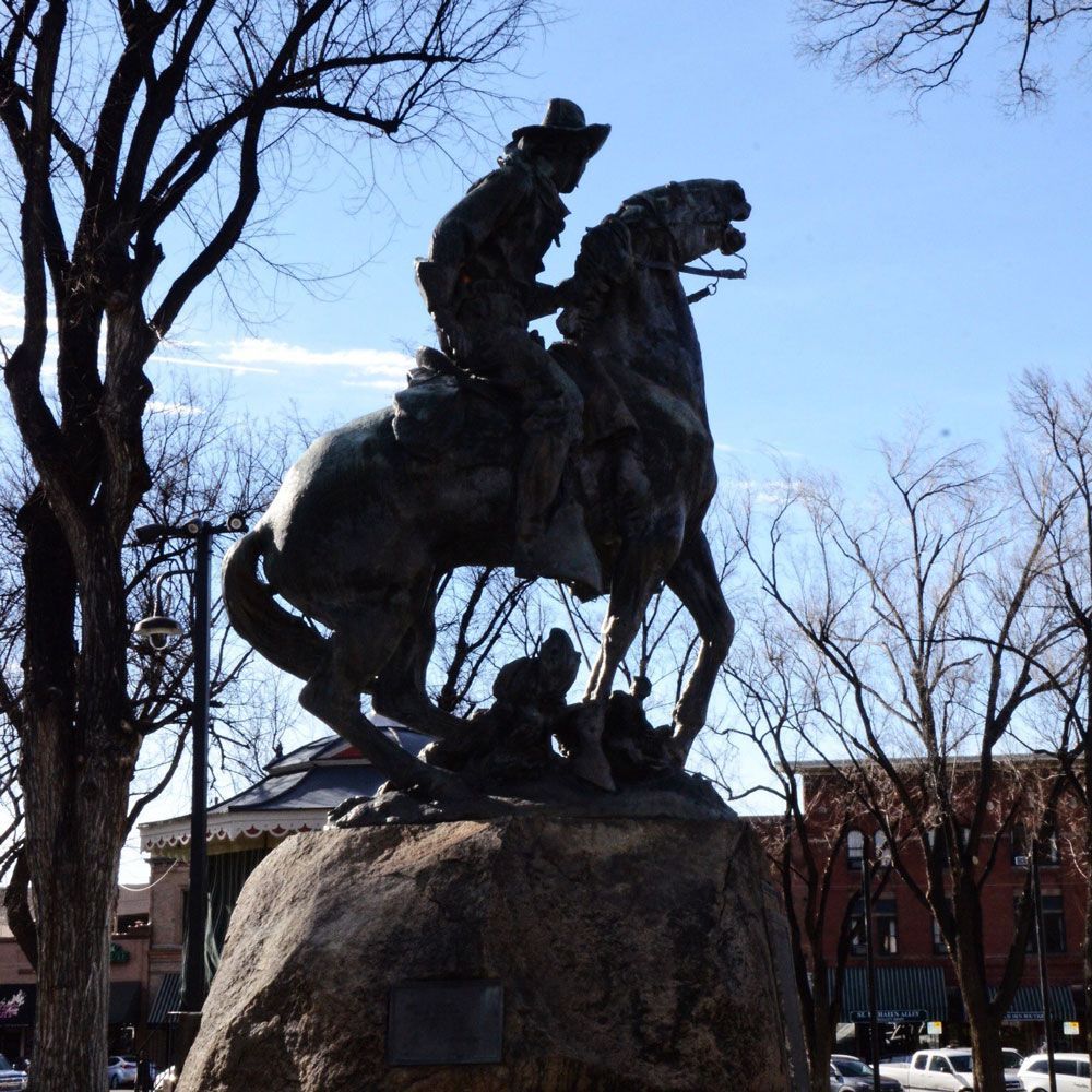 Bronze cowboy statue atop a rock, riding a rearing horse; bare trees, buildings in background.