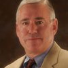 Man with short gray hair, wearing a tan blazer, blue shirt, and maroon tie.