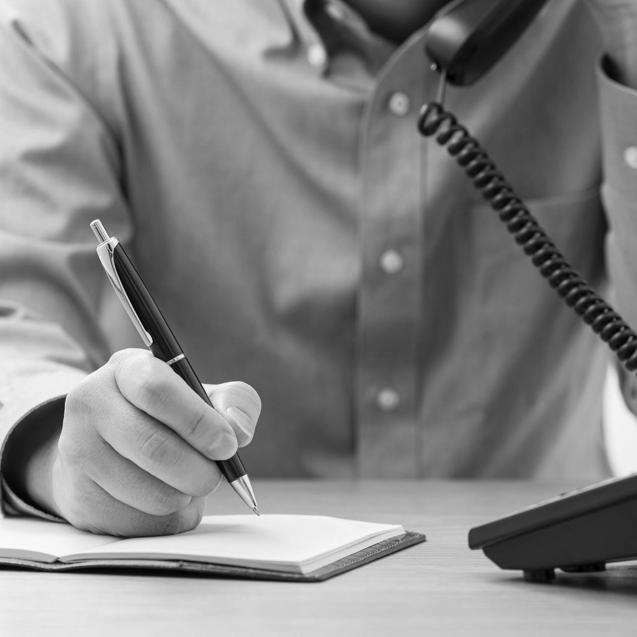 Person writing in a notebook beside a desk phone, with a coiled cord visible.