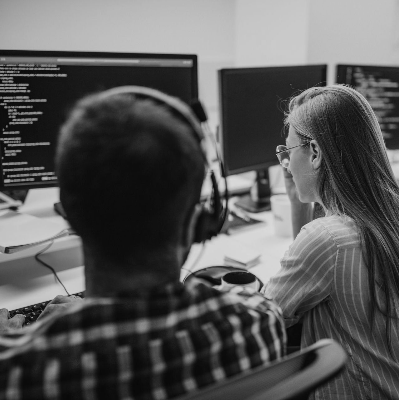 Two coworkers view code on desktop monitors in a shared office, seen from behind