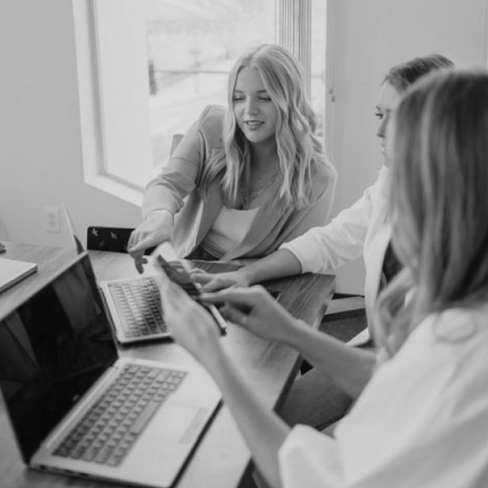 Coworkers collaborating around a laptop, one woman smiling and pointing at the screen in a bright office