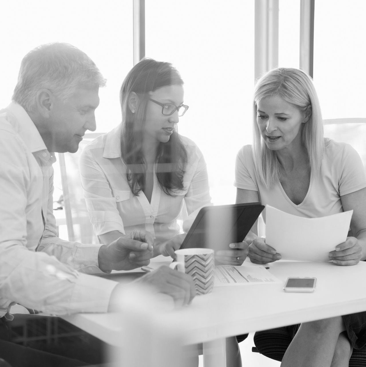 Three coworkers in a bright meeting room reviewing documents around a laptop and coffee cups