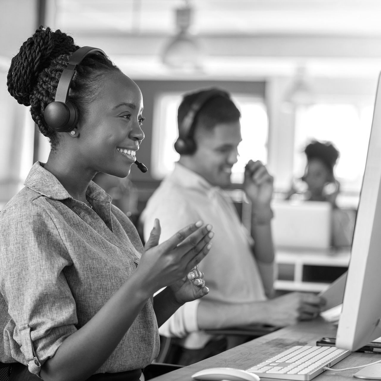 Woman wearing headset claps at desktop computer in an office, with coworkers blurred in the background