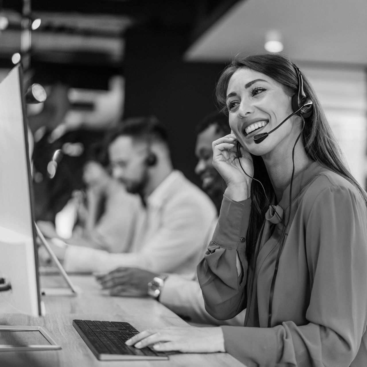 Smiling call center agent wearing a headset, typing at a desk in an office with colleagues blurred in the background