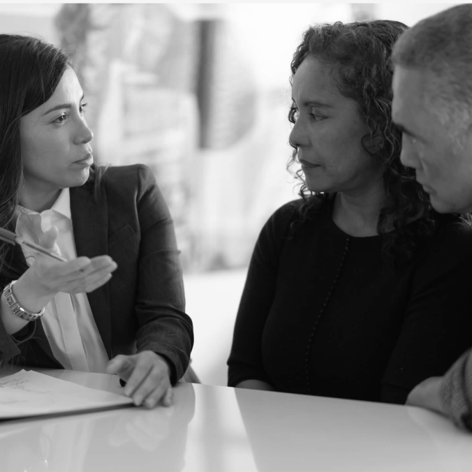 Three people in a meeting, listening and talking around a table in a bright office.