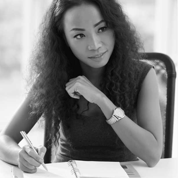 Woman writing in a notebook at a desk, wearing a watch and looking thoughtfully to the side.