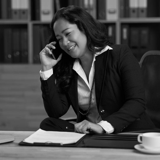 Smiling woman in a black blazer talking on a phone at a desk with an open notebook and coffee cup