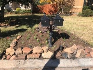 A dual mailbox stand sits in a garden bed bordered by decorative rocks near a curb on a sunny day.