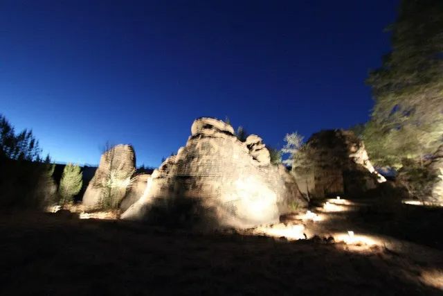 Large rock formations at night, illuminated by soft ground lighting against a deep blue sky.