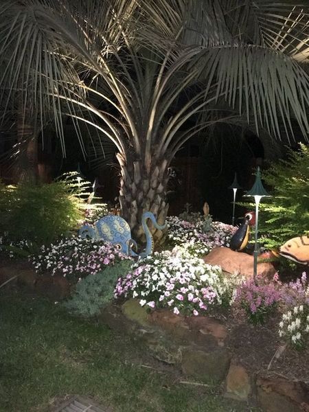 A night scene of a landscaped garden with a central palm tree, glowing white flowers, and whimsical garden sculptures.