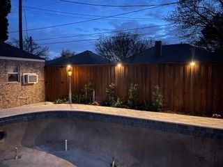 An empty backyard swimming pool at dusk, illuminated by three fence-mounted lights and a pathway light.