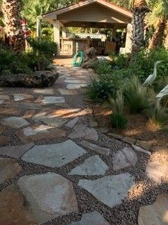 A stone paver path leads through a lush garden toward a covered outdoor patio with a seating area.
