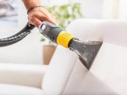Una persona utiliza una máquina limpiadora de tapicería portátil para limpiar un sofá de color claro.