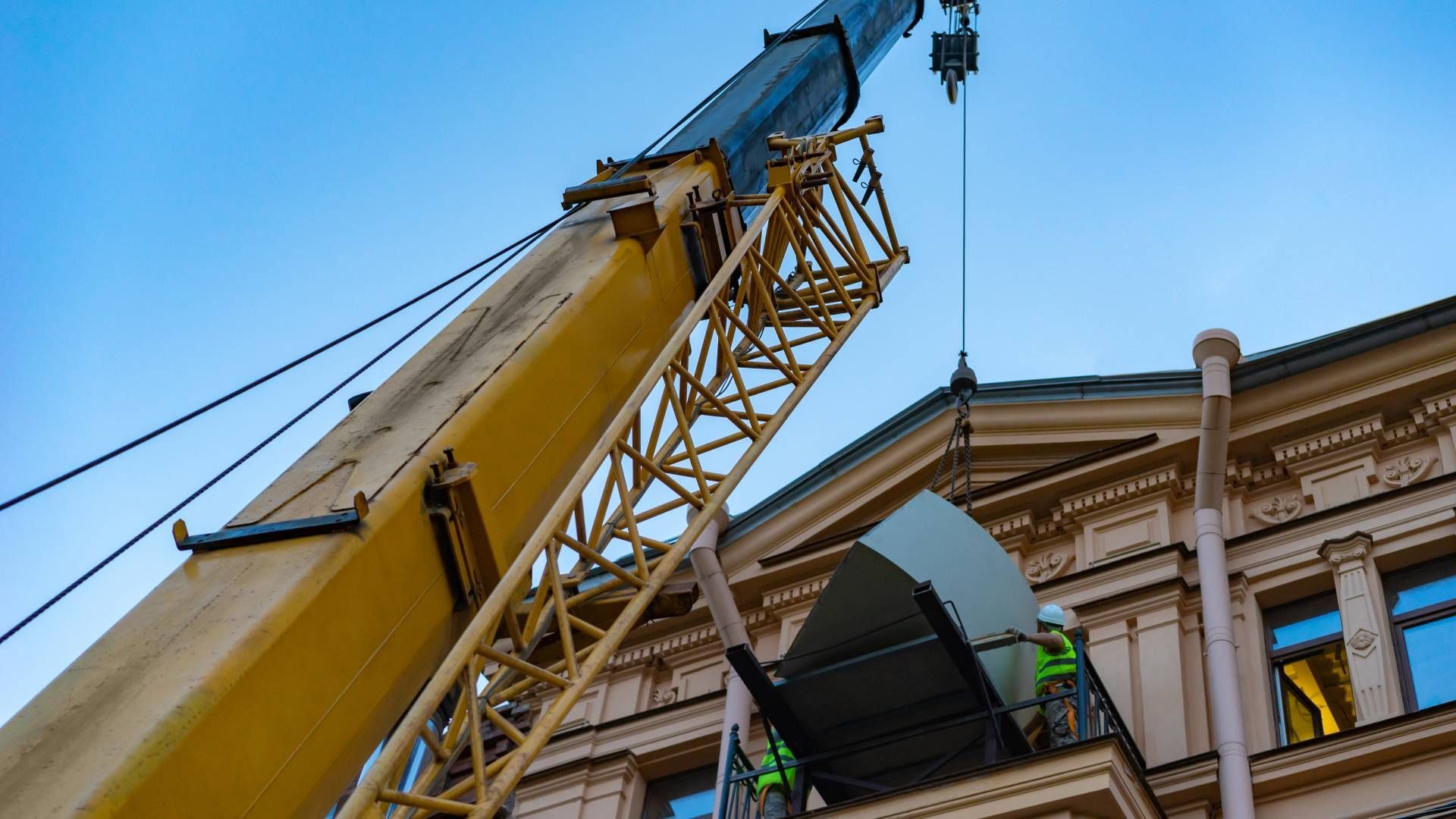 A crane lifting a large object onto a building's roof, workers visible, yellow crane, blue sky.