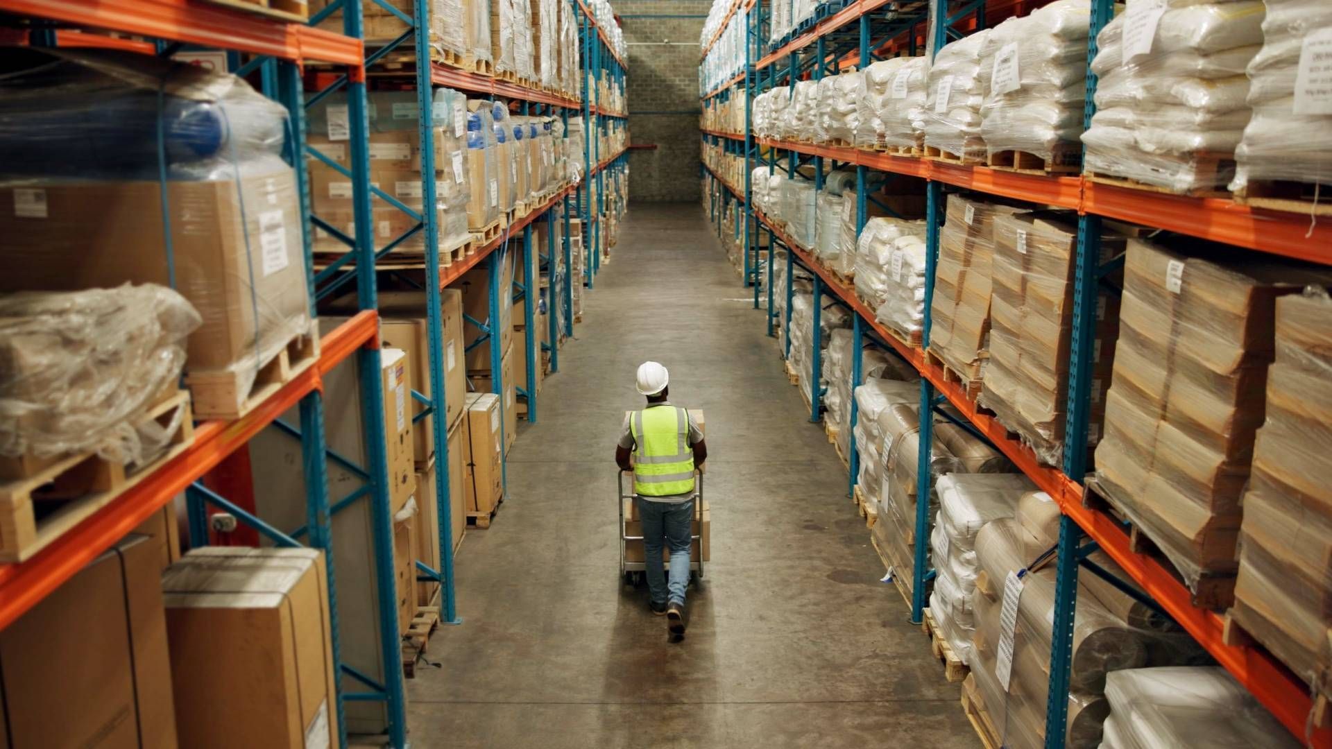 Warehouse worker in safety vest pulls a cart between rows of stocked shelves.