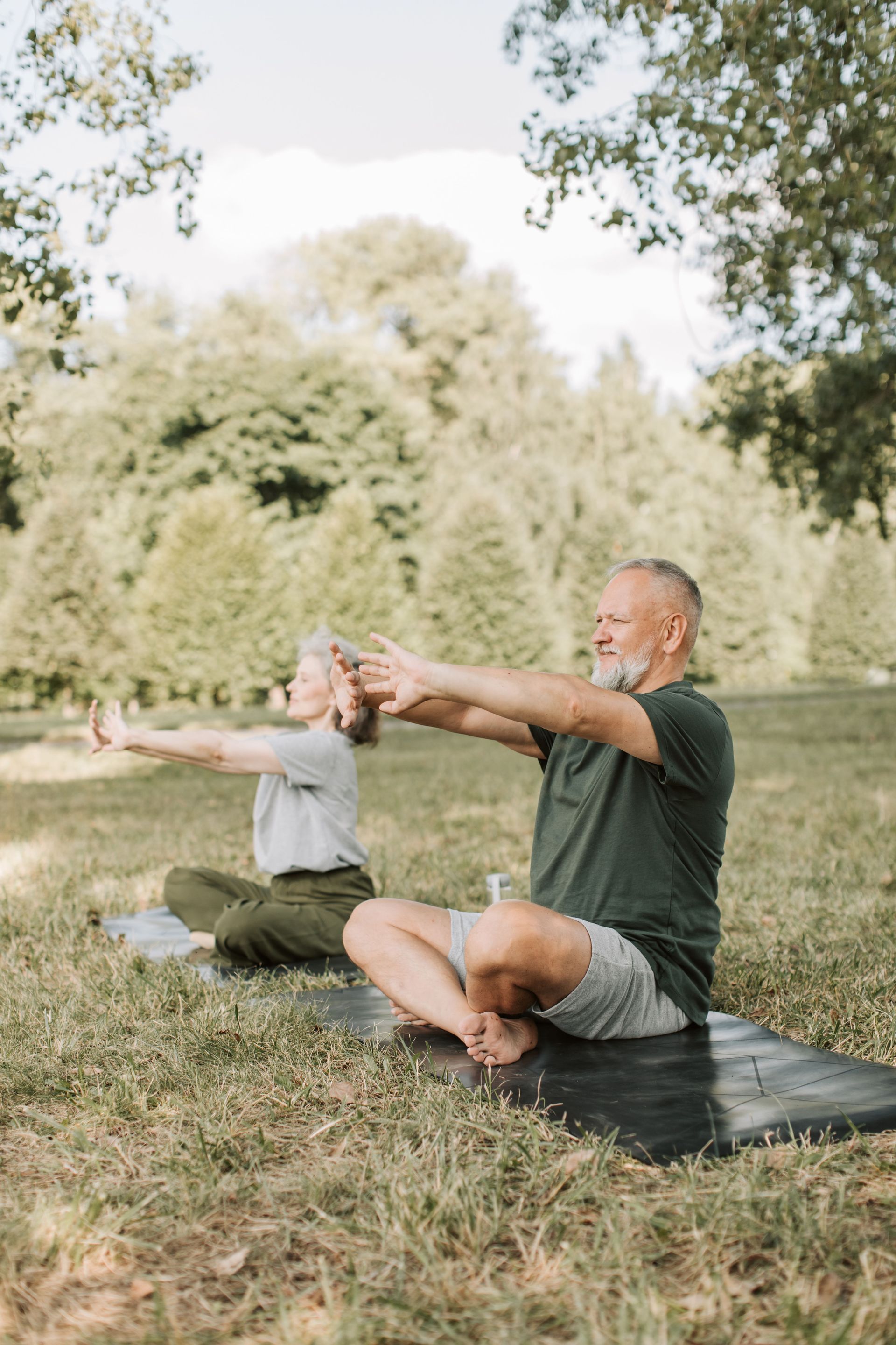 Two people seated on yoga mats stretching outdoors in a grassy park