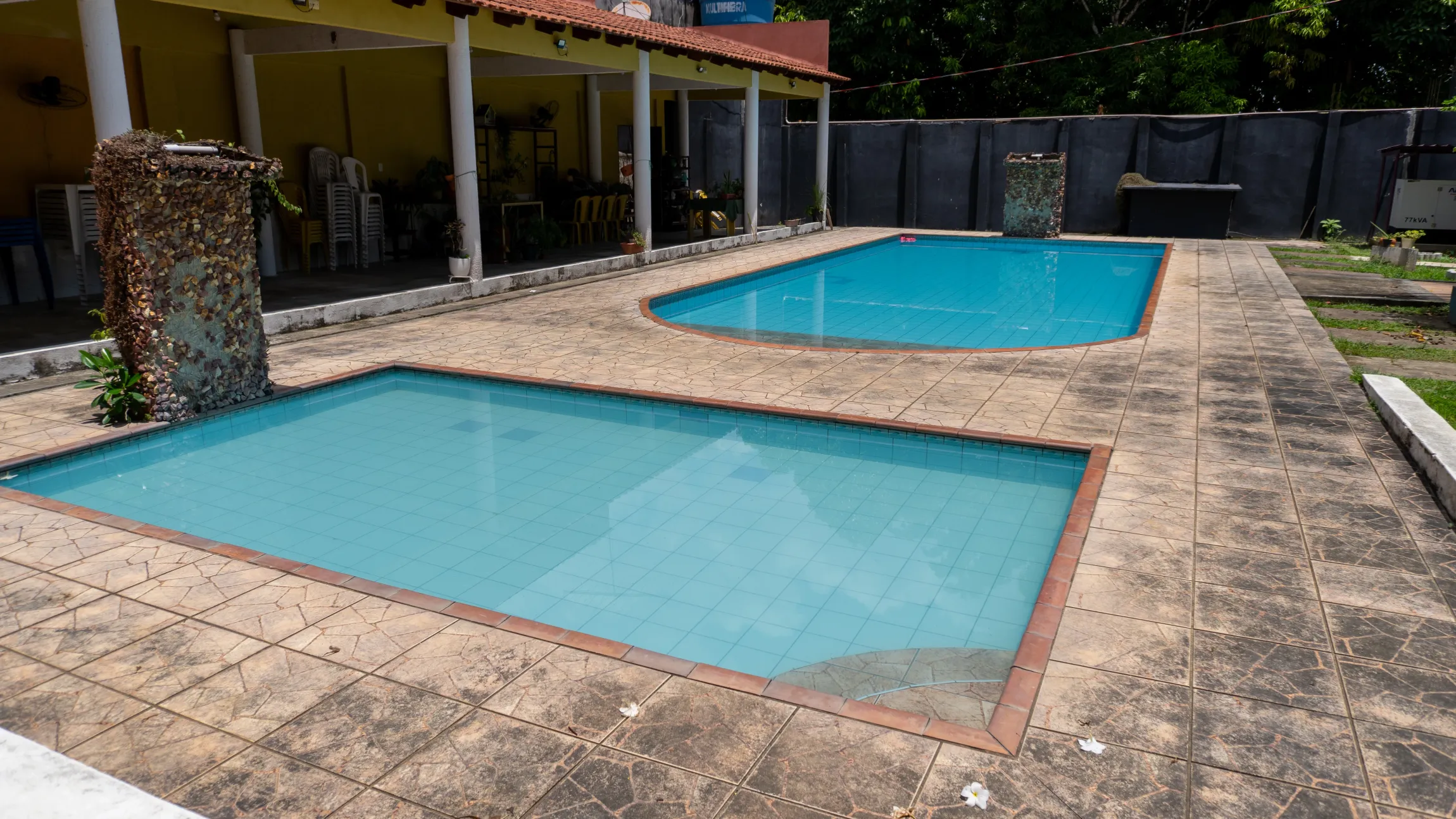 Duas piscinas azuis com pátio de concreto, perto de um prédio amarelo e uma cerca - Solar Café e Pousada