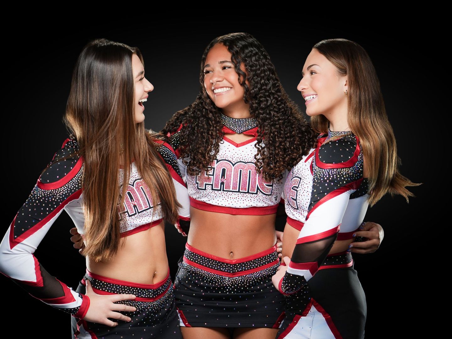 Three cheerleaders in black, red, and white uniforms smiling with arms around each other against a dark background