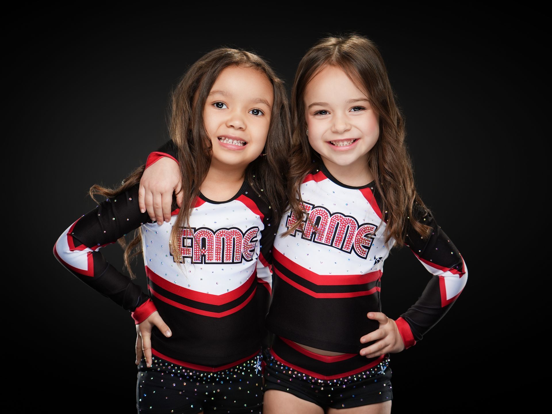 Two cheerleaders in black, red, and white uniforms smiling with arms around each other on a dark background