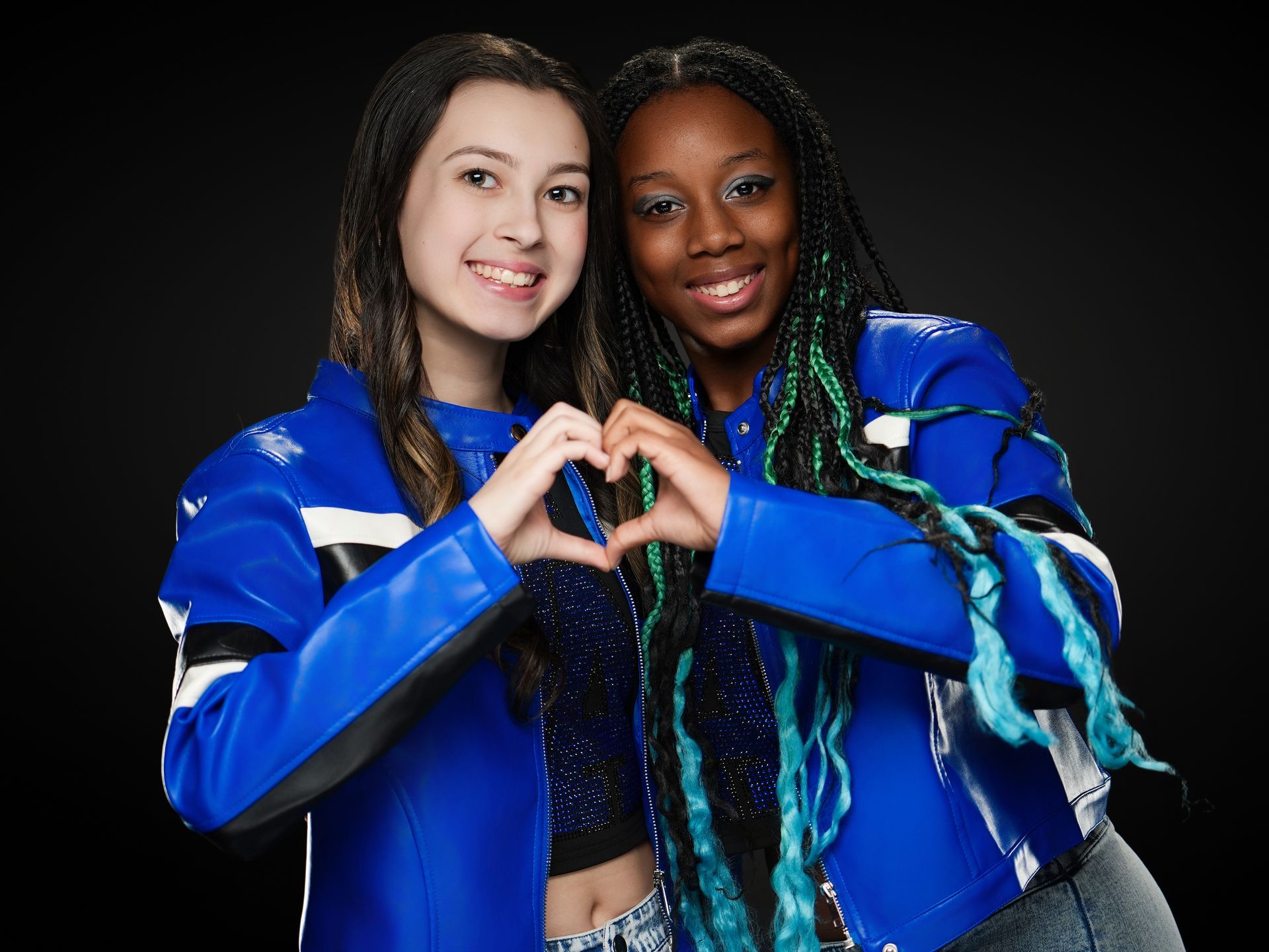 Two women in blue jackets smiling and making a heart shape with their hands against a black background
