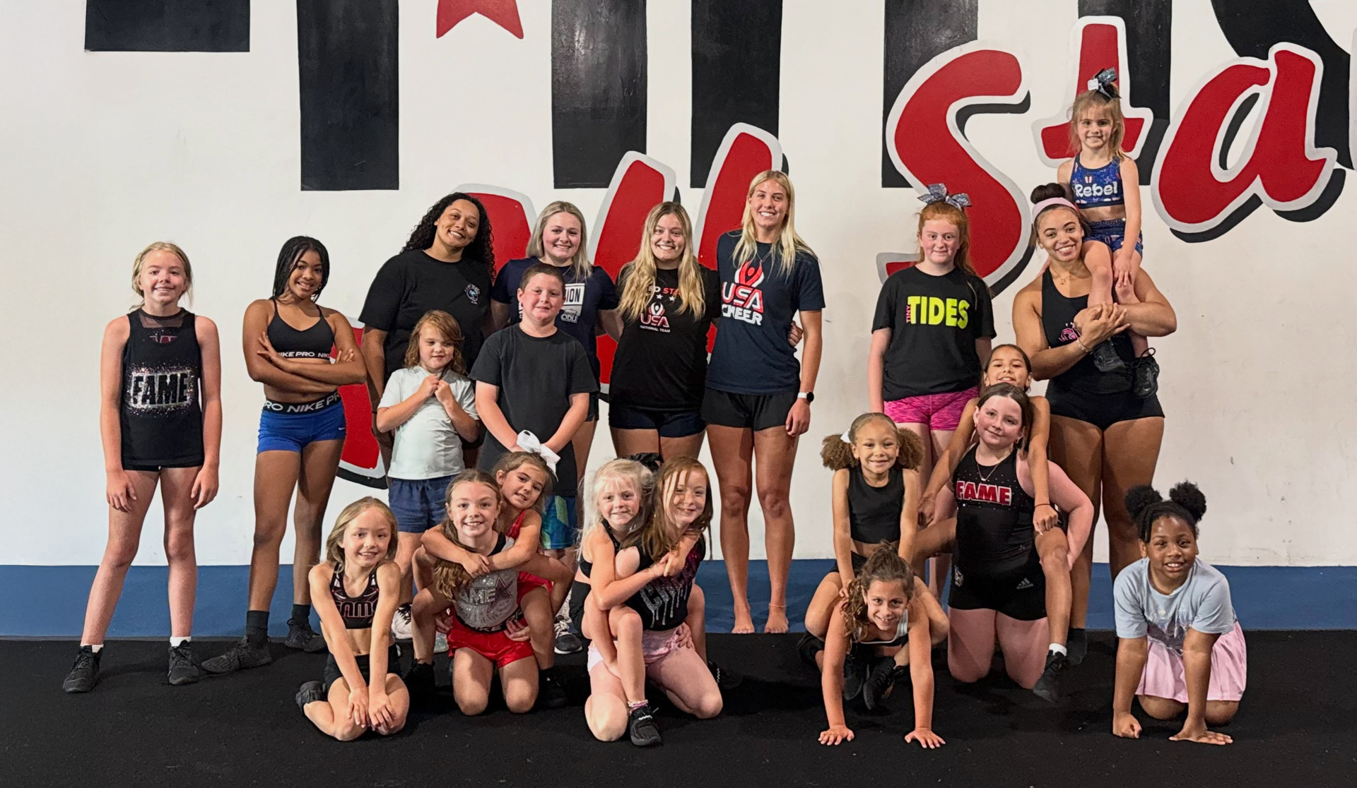 Group of cheerleaders posing in a gym, wearing black and red uniforms with red pom-poms.