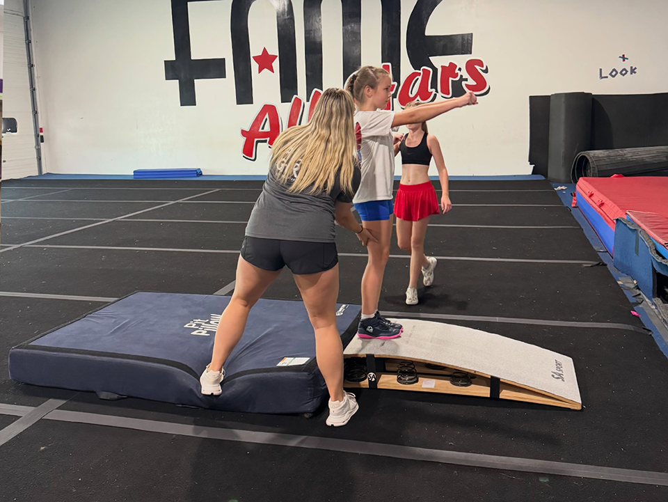 Three gymnasts on a spring floor near mats, with one balancing on a tilted board in an All Stars gym.