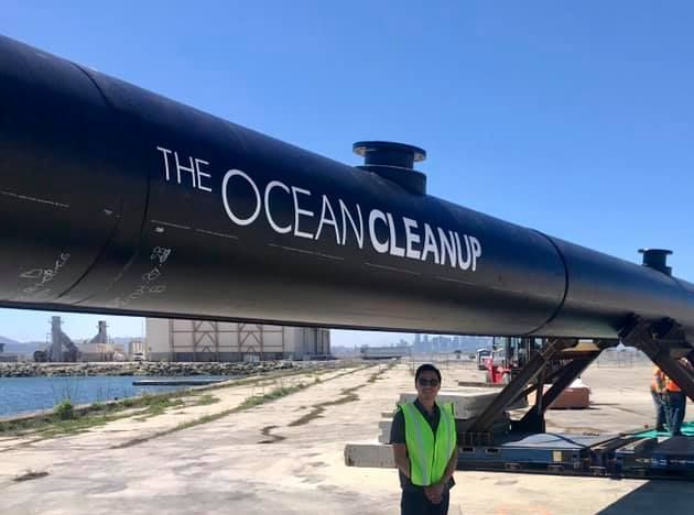A man in a yellow vest is standing in front of a large black pipe that says the ocean cleanup.