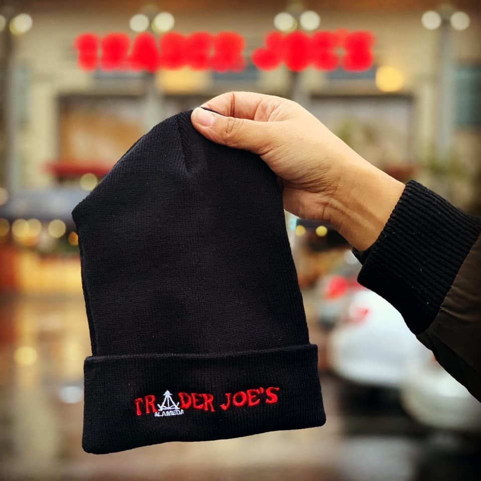 A person is holding a black beanie that says trader joe 's