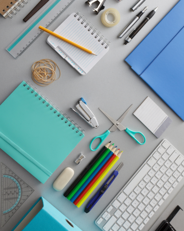 Composizione piatta di colorati articoli di cancelleria e materiale scolastico su una scrivania grigia, tra cui quaderno, forbici, penne, righello e tastiera.