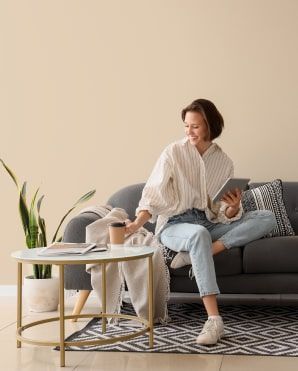 Una donna seduta su un divano grigio, con in mano un tablet e sorridente, è seduta accanto a un tavolino da caffè in un accogliente soggiorno.