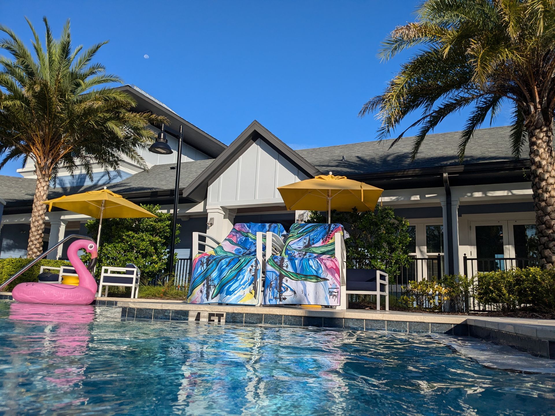 two beach and or yoga towels draped over two chairs that sit next to the pool. there are yellow beach umbrellas behind the colorful towels. There is a pink flamingo float in the pool in front of a house.there is a canary island date palm tree to the right of the beach towels.