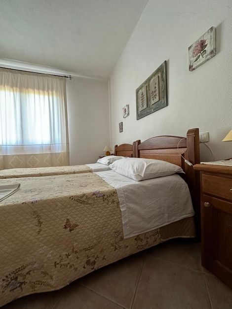 Piccola camera da letto con letto in legno, copriletto a fantasia, comodino e finestra con tende trasparenti.