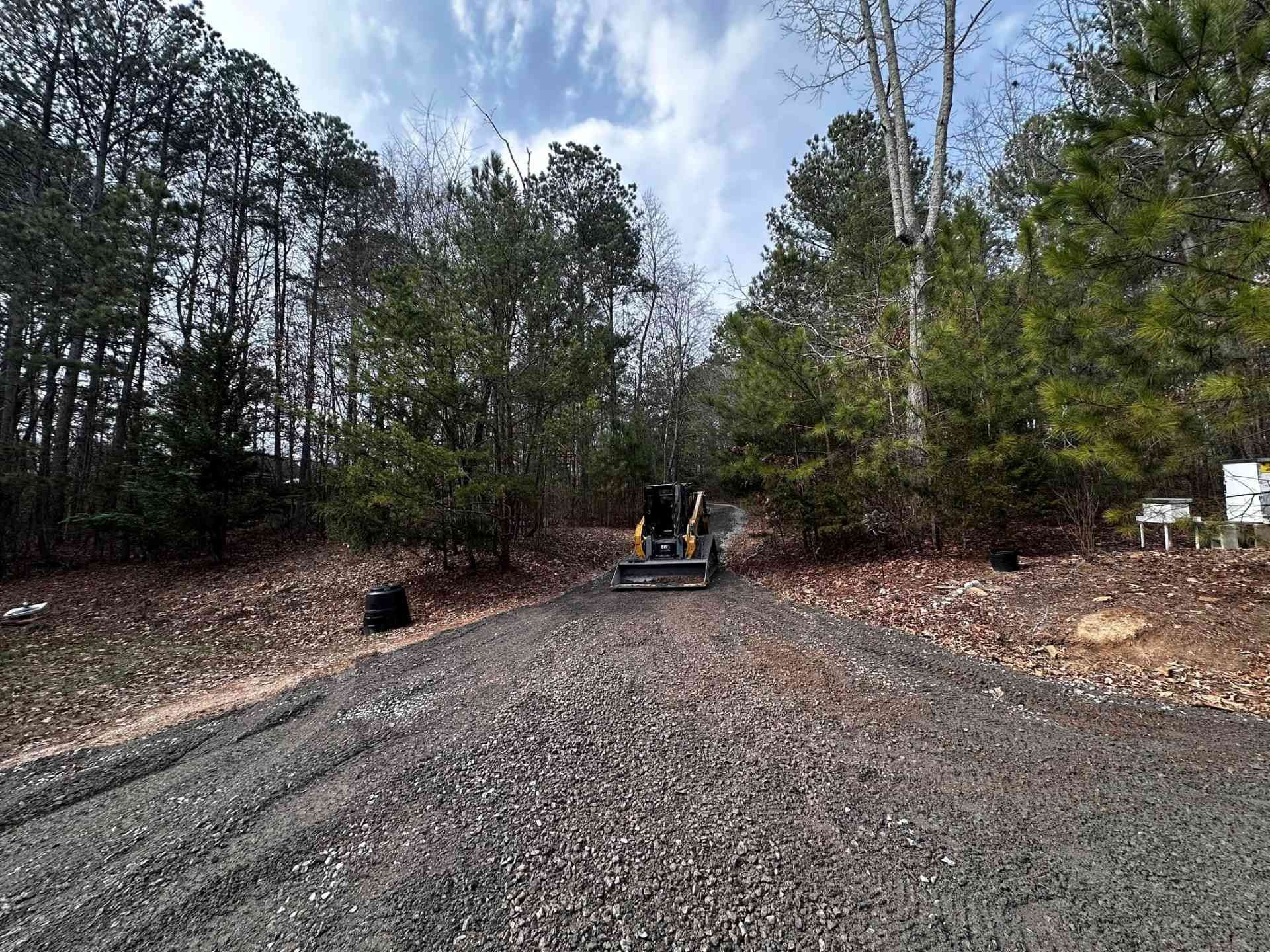 A construction vehicle sits on a freshly laid gravel driveway leading into a dense, wooded forest area under a blue sky.