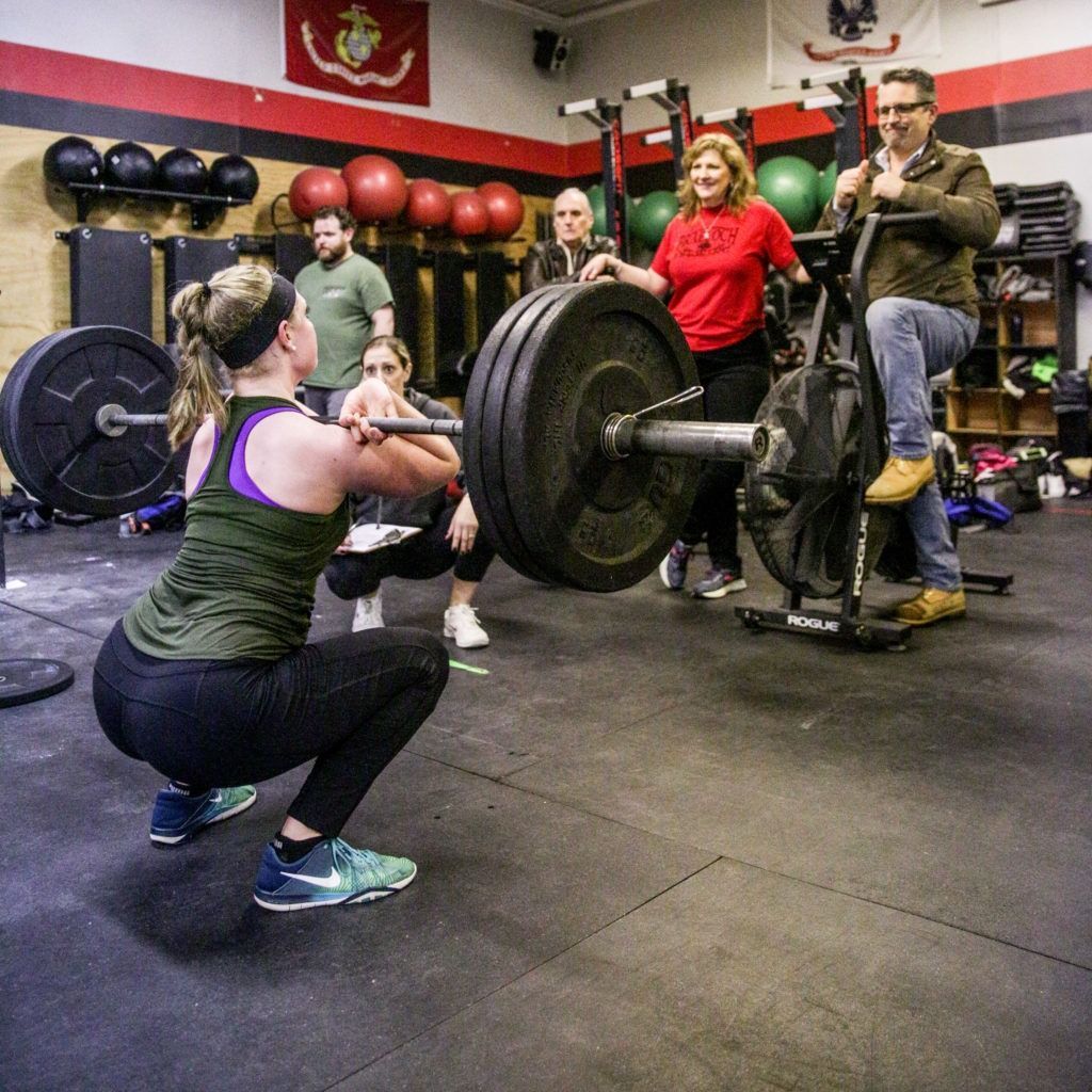 A woman is squatting down with a barbell in a gym while a group of people watch.