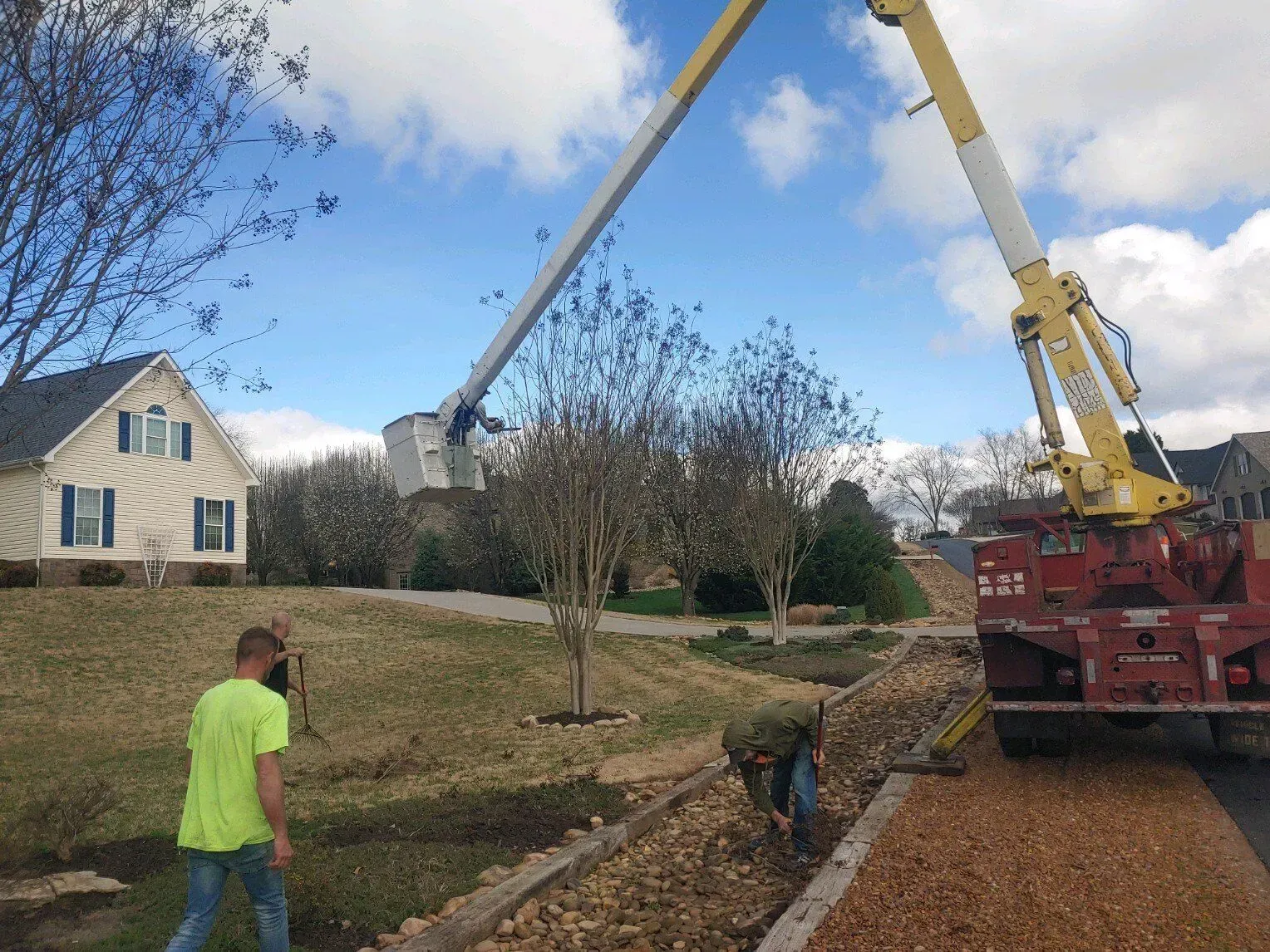 Worker and crane truck clearing a tree-lined roadside beside a house on a suburban street