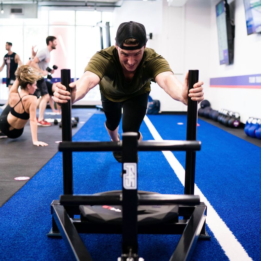 Person pushing a weighted sled on a blue gym turf track, training in a fitness facility.